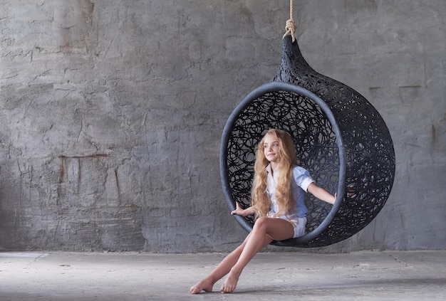 Cute young woman posing near big oval black artistic chair over grey background.