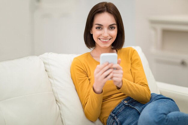 Милая молодая женщина отдыхает дома на диване с помощью смартфона