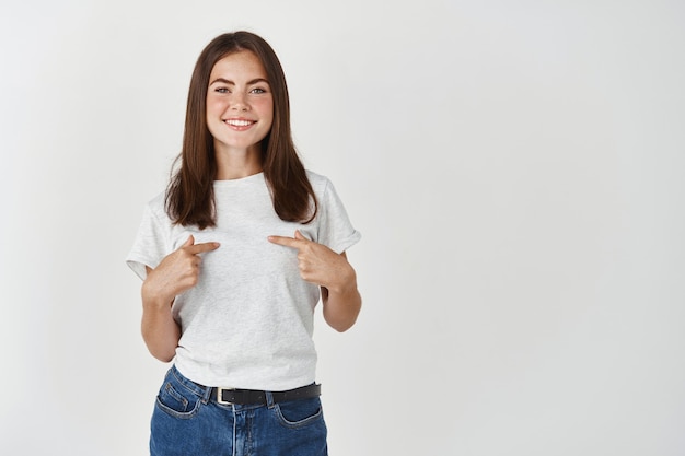 Cute young woman smiling and pointing at herself, you need me gesture, standing over white wall