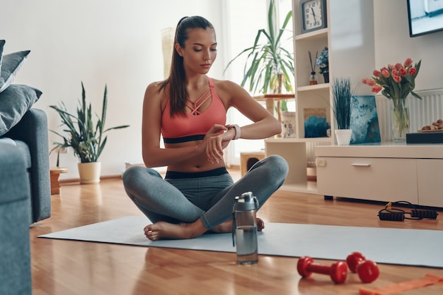 Cute young woman in sports clothing checking her heart rate using smart watch while sitting in lotus position at home