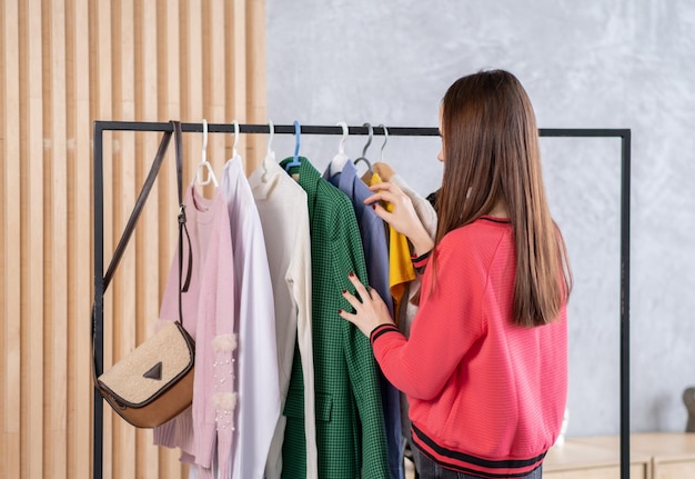 Cute young woman standing in front of hanger rack and trying to choose outfit dressing for work or walk Selection of a wardrobe stylist shopping