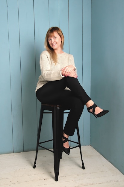 Cute young woman with blond flowing hair just below her shoulders, sitting on high chair legs crossed on wall of blue wooden boards.