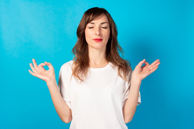 Cute young woman with closed eyes and a white t-shirt makes a gesture with his hands yoga, concentration, meditation on isolated blue. Concept of meditation, dreams, planning, good mood