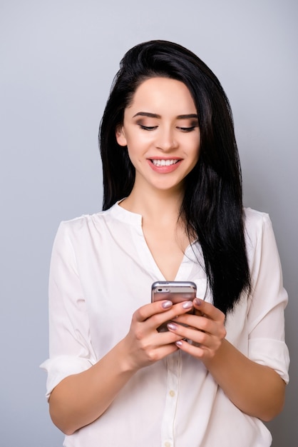 Cute young woman with thick black hair read sms from her boyfriend with toothy smile while standing on gray space