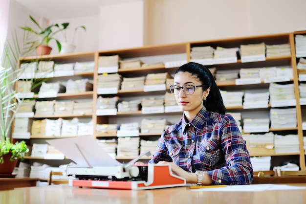 Lo scrittore sveglio della giovane donna in vetri sta scrivendo su una macchina da scrivere nella biblioteca di fondo