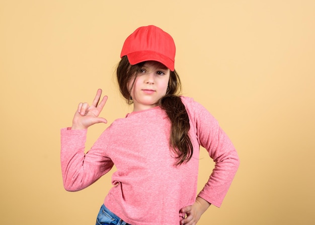 Cutie in cap Girl cute child wear cap or snapback hat beige background Little girl wearing bright baseball cap Modern fashion Kids fashion Stylish accessory Feeling confident with this cap