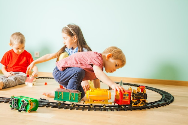 Симпатичный маленький мальчик играет с игрушечной железной дорогой на полу перед своими братом и сестрой