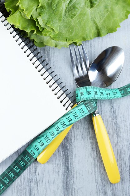 Cutlery tied with measuring tape and notebook with lettuce on wooden background