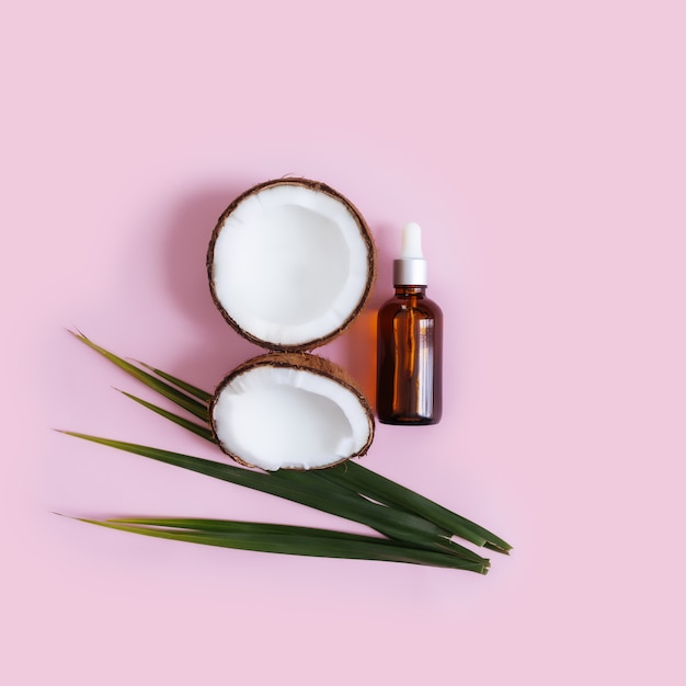 Cuts and halves of coconut and green palm leaf, glass bottle with essential oil on a pastel pink background.