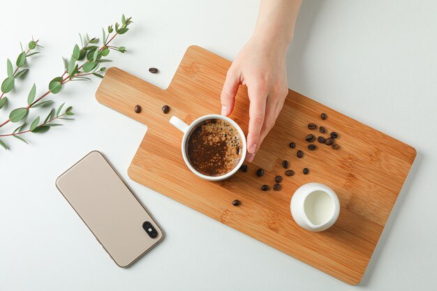 Cutting board with cup of coffee, coffee beans, phone and plant branch on white background