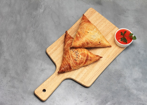 Cutting board with delicious meat samosas and red sauce on gray