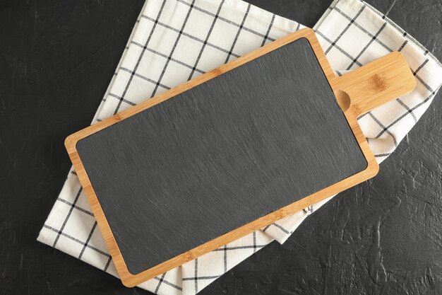 Cutting board with kitchen towel on black background, top view