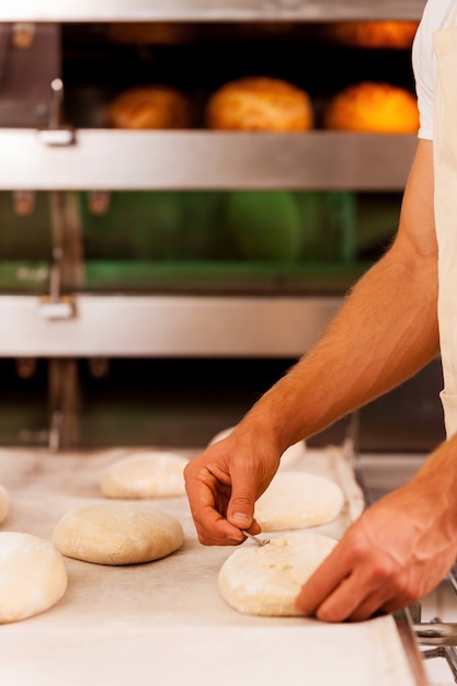 Cutting the dough. Close-up of man cutting the dough while standing against oven with bread in it