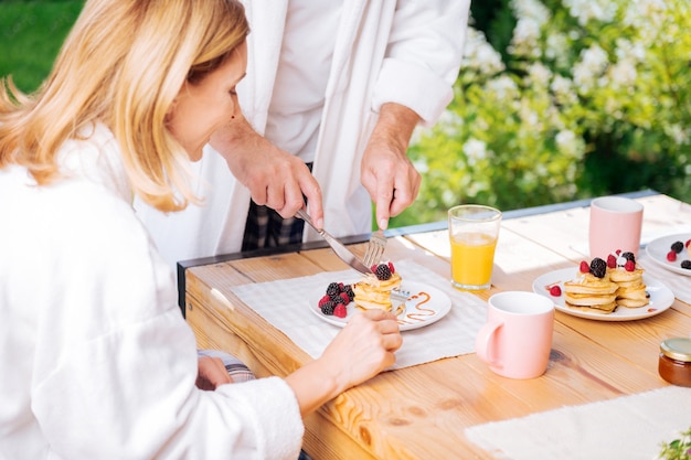 Cutting pancakes. Loving husband wearing pajama and bathrobe cutting pancakes for his beautiful blonde-haired wife