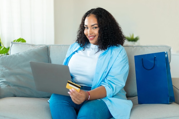 Cybershopping concept Excited black chubby lady with laptop and credit card ordering things online at home