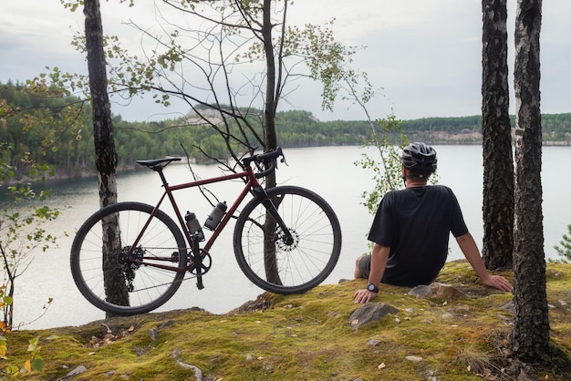 Il ciclista in un casco siede vicino alla sua bicicletta sul bordo di una collina vicino a un pittoresco lago