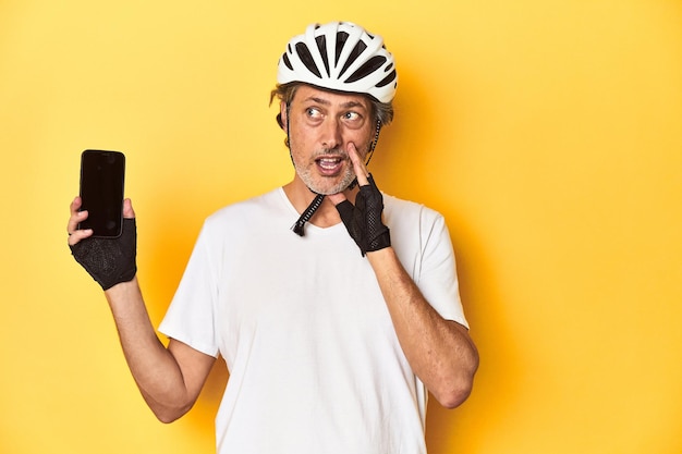 Cyclist man showing phone on yellow backdrop is saying a secret hot braking news and looking aside