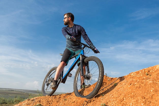 Cyclist in shorts and jersey on a modern carbon hardtail bike with an air suspension fork standing on a cliff against the background of fresh green spring forest