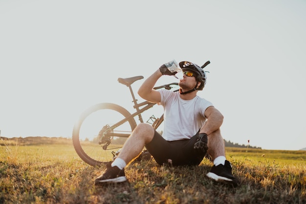 산악 도로에서 격렬한 라이딩에서 휴식을 취하는 동안 자전거 타는 사람은 초원에 앉아 있습니다. 자전거 타는 사람은 물로 냉각됩니다. 선택적 초점 고품질 사진