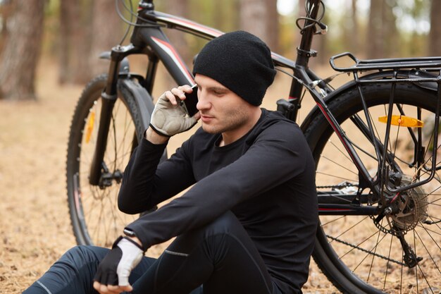 Cyclist sitting on ground, holding smartphone in one hand, having conversation over his mobile phone, leaning on bicycle, relaxing alone around nature, taking break to have rest.