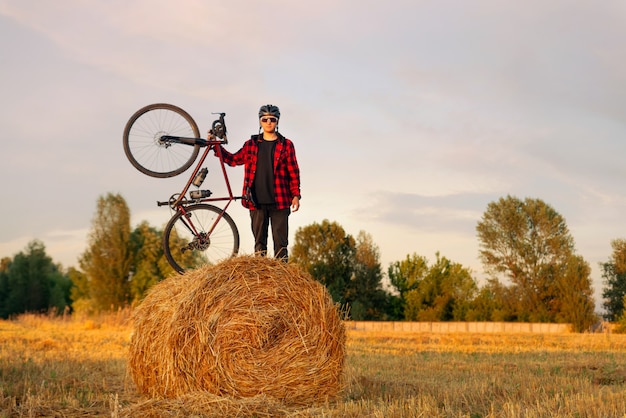 Cyclist with gravel bicycle stands on top of a haystack in a field at sunset.