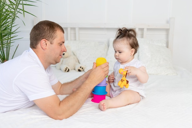 Dad and baby girl play with colorful toys on the bed at home in a bright room