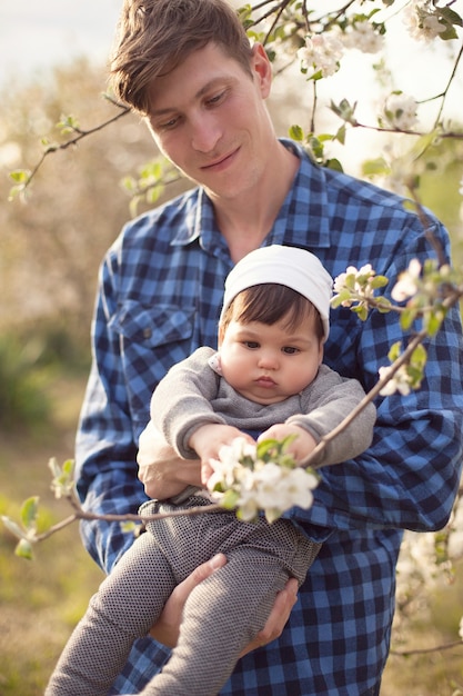 Dad in a check shirt holds his little son in his arms and looks at the apple tree flowers against the backdrop