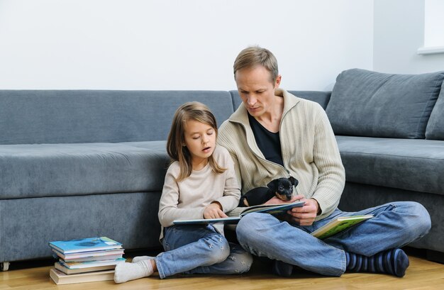 Dad and daughter are reading books. The puppy is lying on the man's legs.