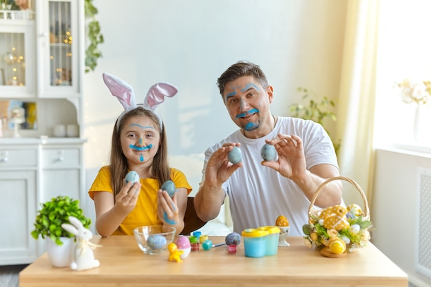 Dad and daughter faces stained with blue paint for painting eggs. on the table is a basket with Easter eggs and paints.