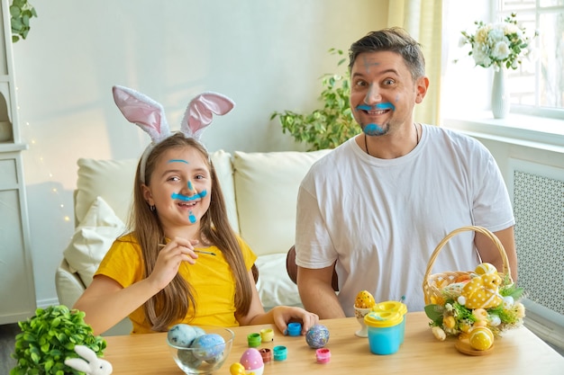 Dad and daughter faces stained with blue paint for painting eggs. on the table is a basket with Easter eggs and paints.