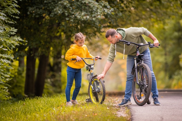 Papà e figlia ispezionano la ruota della bicicletta adolescente per bambini sul sentiero autunnale del parco.