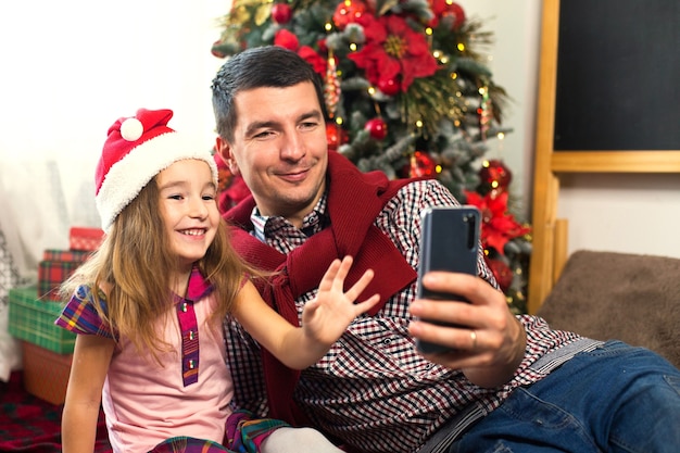 Dad and daughter near the Christmas tree with a smartphone take a selfie, communicate via video connection. Christmas greetings, a gift box, a girl in a Santa hat waving Hello. New year, holiday decor