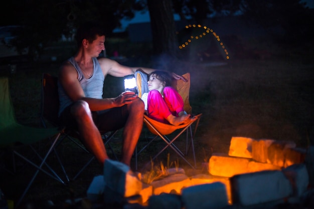 아빠와 딸은 여름에 자연 속에서 야외에서 난로 옆에 밤에 앉아 있습니다. 가족 캠핑 여행, 모닥불 주변 모임. 아버지의 날, 바베큐. 캠핑 랜턴과 텐트