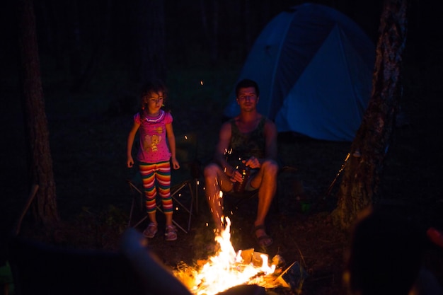 お父さんと娘は自然の中で夏の野外で火のそばに夜座っています。家族でのキャンプ旅行、キャンプファイヤーの周りの集まり。父の日、バーベキュー。キャンプ用ランタンとテント