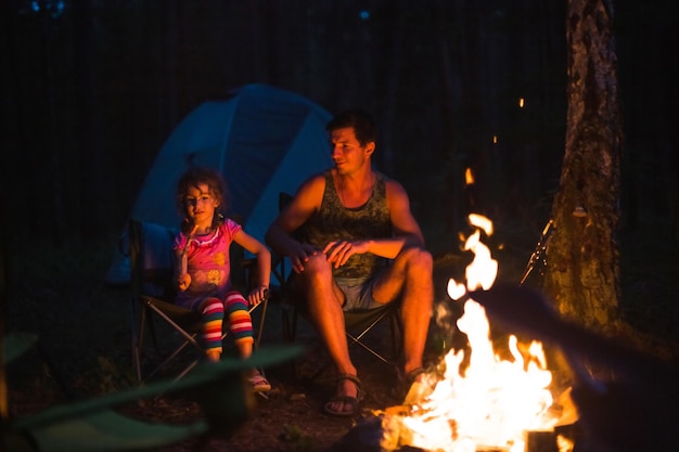 Papà e figlia si siedono di notte accanto al fuoco all'aria aperta in estate nella natura viaggio in famiglia in campeggio riunioni intorno al fuoco barbecue festa del papà lanterna e tenda da campeggio