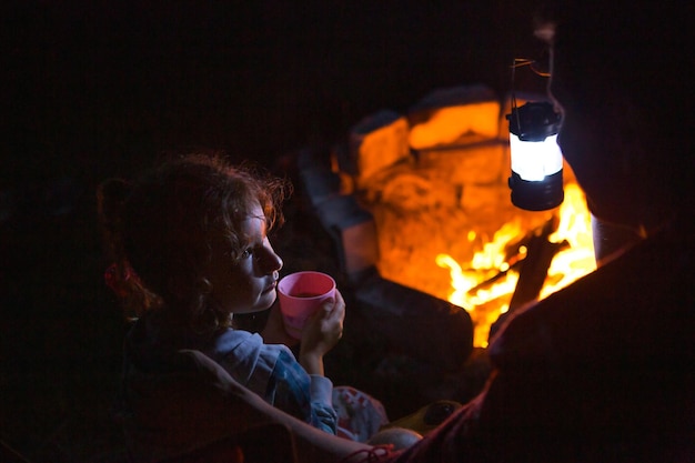 Dad and daughter sit at night by the fire in the open air in the summer in nature Family camping trip gatherings around the campfire Father's Day barbecue Camping lantern and tent