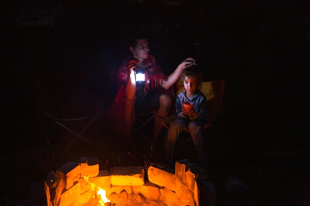 Dad and daughter sit at night by the fire in the open air in\
the summer in nature family camping trip gatherings around the\
campfire father\'s day barbecue camping lantern and tent