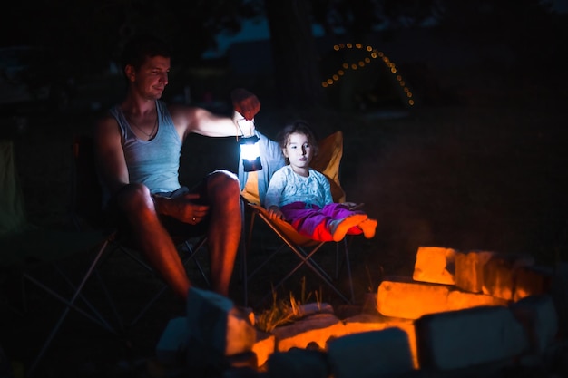 Dad and daughter sit at night by the fire in the open air in\
the summer in nature family camping trip gatherings around the\
campfire father\'s day barbecue camping lantern and tent