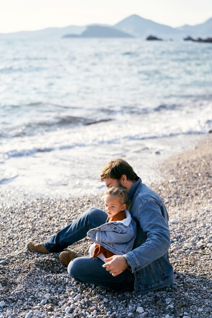 Dad in a denim jacket sits on a pebble beach and holds a little girl in his lap side view