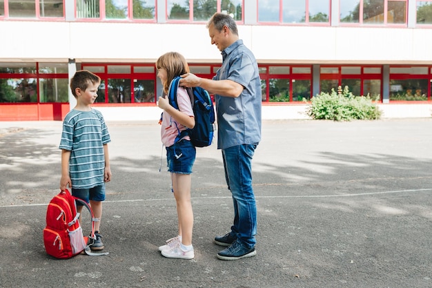 写真 父親は子供たちを学校に連れて行き ⁇ 娘に女子校生のためにバックパックを着せるのを手伝います ⁇