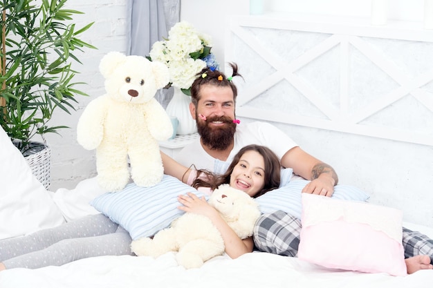 Dad and girl relaxing in bedroom Pajamas style Father bearded man with funny hairstyle ponytails and daughter in pajamas Having fun pajamas party Slumber party Happy fatherhood Close friends