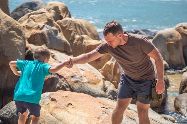 Dad help son jump down over stone walk spend time together man\
hold kid hand sea mountain nature education father day family union\
hiking raising boy influence in happy child live worldview