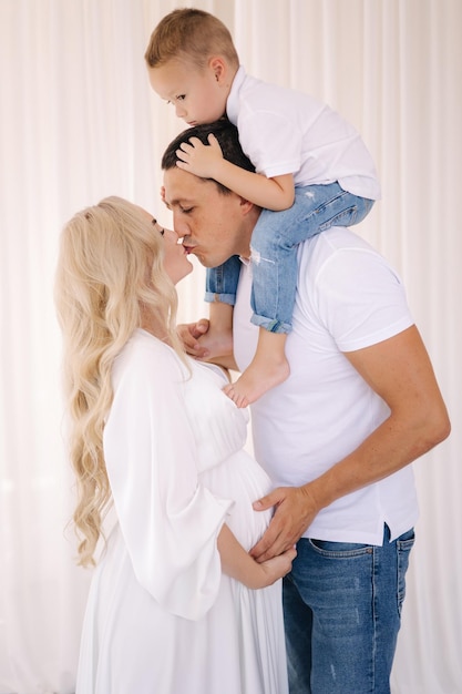 Dad hold his son on neck and hug his pregnat wife family look concept for studio photosession