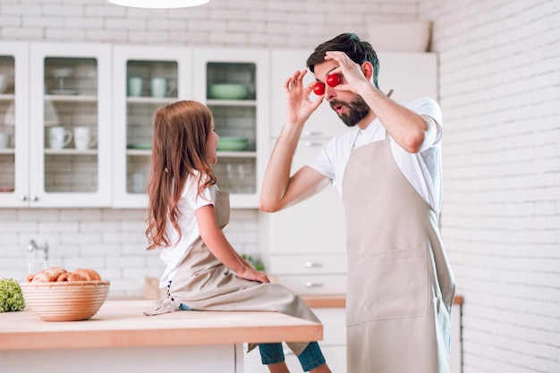 Dad holding cherry tomatoes on his eyes with girl sitting on the table on domestic kitchen