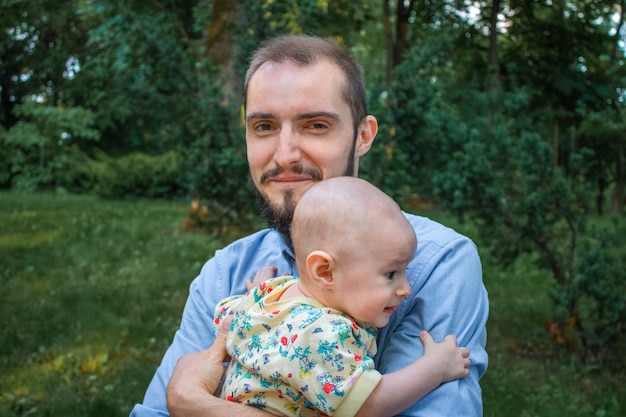 Dad holding his baby son close-up on the background of a summer landscape. Bearded man smiling and happy, looking at camera with confused baby boy