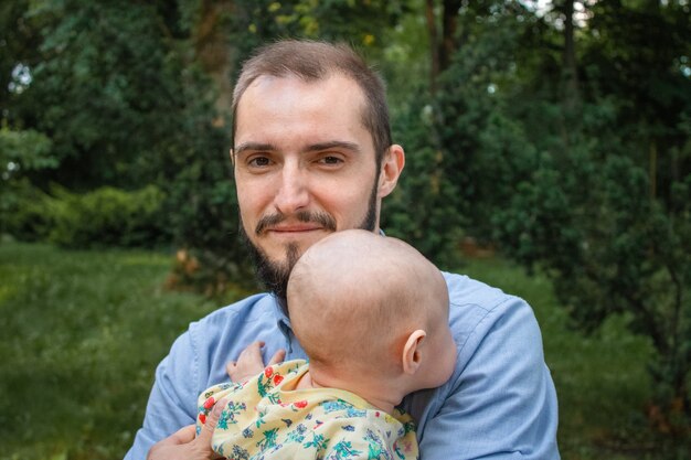 夏の風景の背景に彼の赤ん坊の息子のクローズアップを保持しているお父さん。新生児と一緒にカメラを見て、笑顔で幸せなひげを生やした若い男