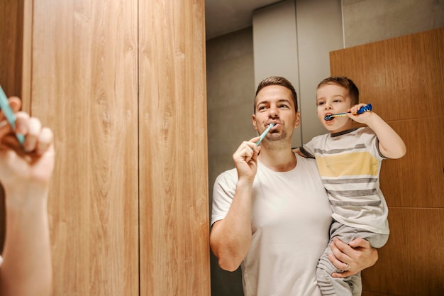 A dad holding his son in the arms while standing in the bathroom and brushing their teeth together
