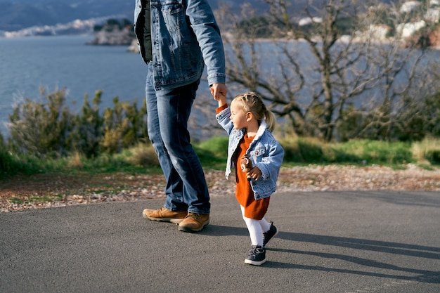 Foto papà tiene la mano di una bambina con un vestito e una giacca di jeans in piedi sulla strada contro il