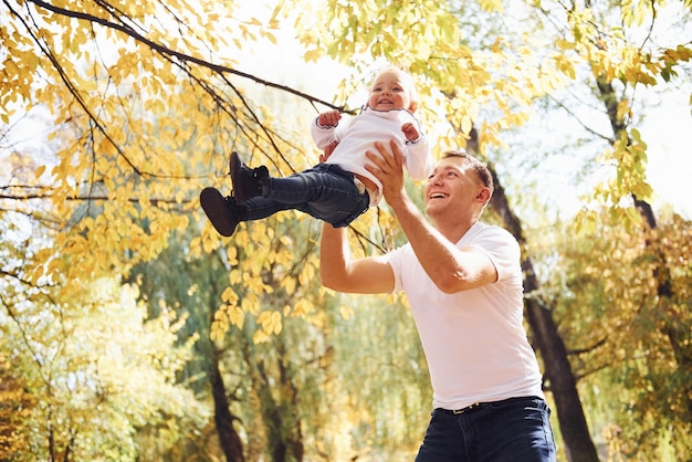 Dad holds his daughter in hands and have fun in the autumn park.