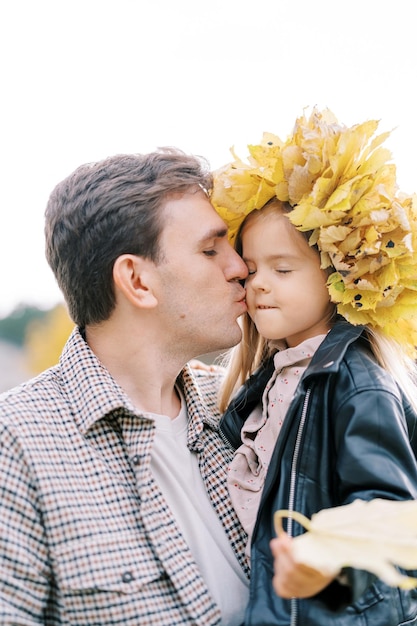 Foto papà bacia la bambina sulla guancia indossando una ghirlanda di foglie gialle tra le braccia ritratto